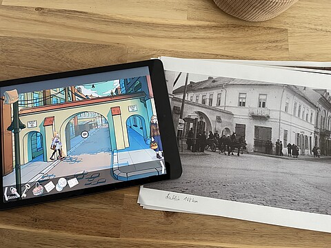 Ein Tablet mit einer Szene aus dem digitalen Spiel liegt neben einem historischen Foto mit einer Straßenszene aus Radom