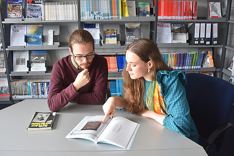 Gabriel and Uliana sitting at a table in the library of the Neuengamme Concentration Camp Memorial and reading a book that is open on the desk